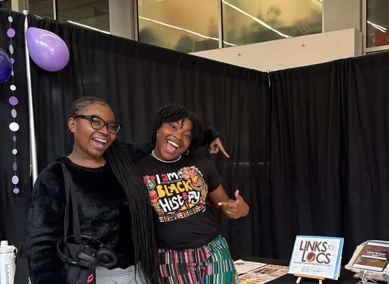 Two women at an event stand beside a table displaying a sign