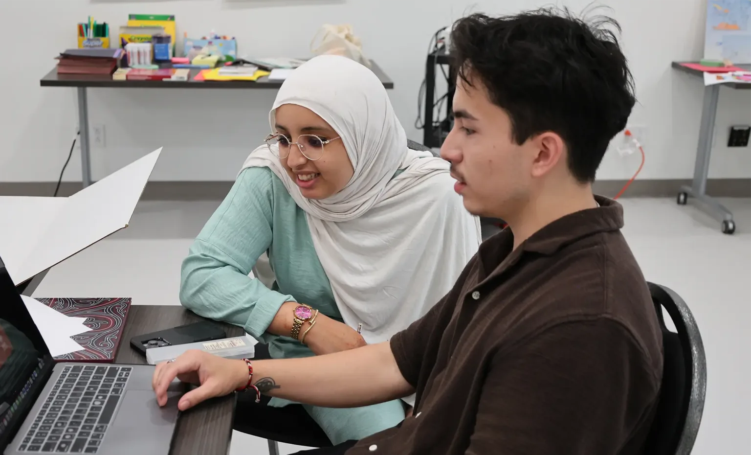 Two individuals collaborate on a laptop in a classroom setting, focused on their work.